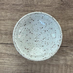 May include: A small, white ceramic bowl with a speckled brown glaze. The bowl is round and shallow, with a slightly flared rim.