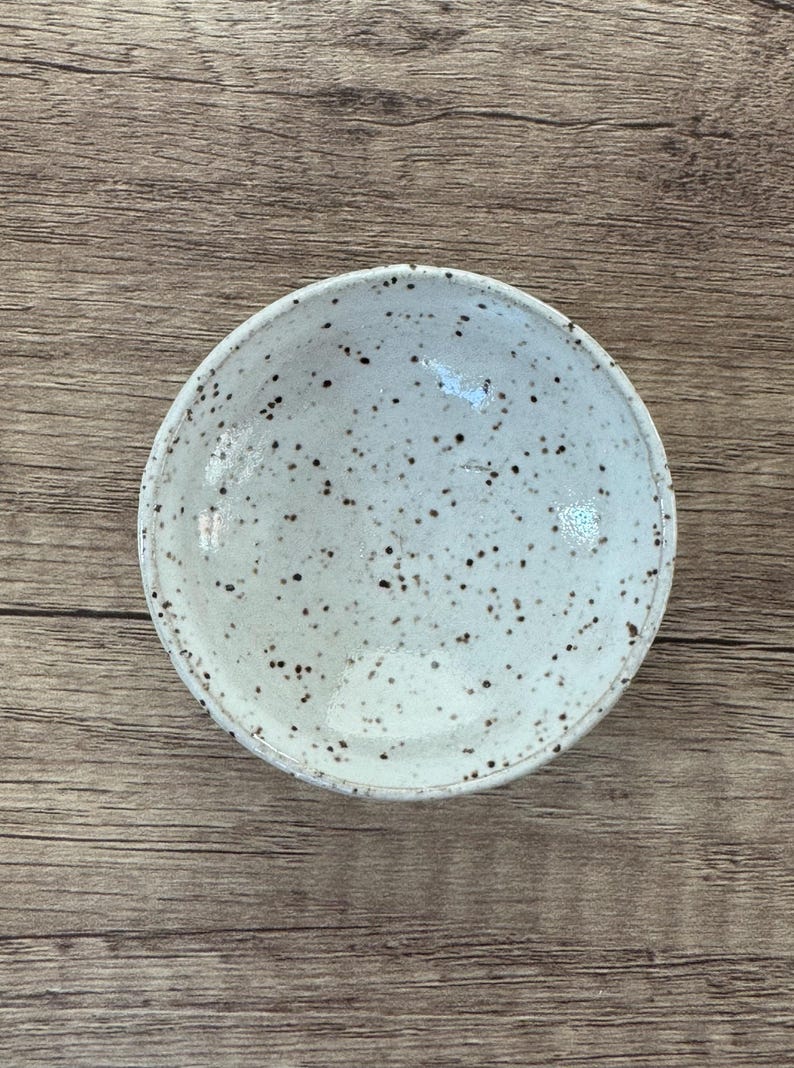 May include: A small, white ceramic bowl with a speckled brown glaze. The bowl is round and shallow, with a slightly flared rim.