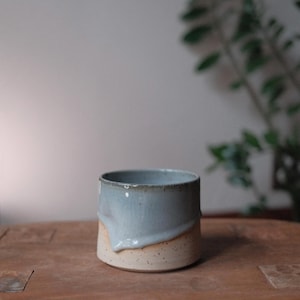 May include: A handmade ceramic mug with a cylindrical shape. The top half is a light blue glaze that drips down over the speckled beige base. The mug is sitting on a wooden surface.