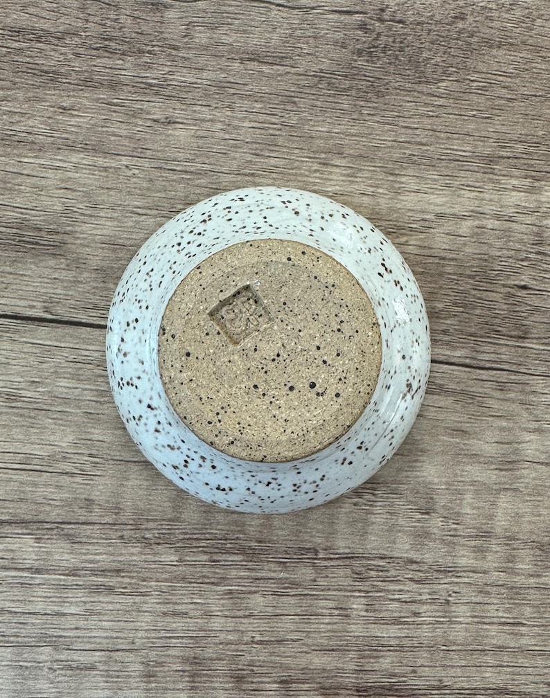 May include: White ceramic bowl with speckled brown glaze. The bowl has a small square indentation on the bottom.