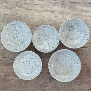 May include: Five small white ceramic bowls with a speckled black and white design. The bowls are arranged in a semi-circle on a wooden surface.