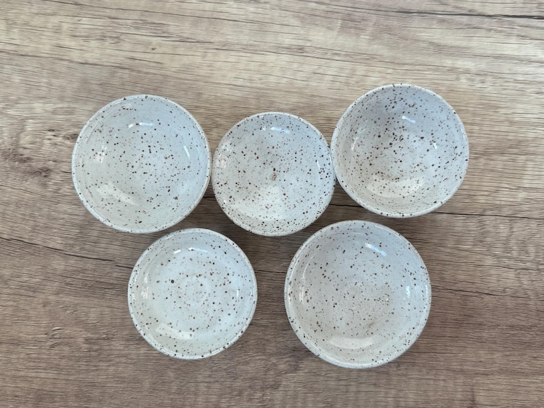 May include: Five small white ceramic bowls with a speckled black and white design. The bowls are arranged in a semi-circle on a wooden surface.