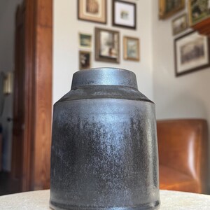 May include: A dark gray ceramic vase with a cylindrical body and a slightly flared top. The vase has a textured surface and sits on a light-colored surface. The background includes framed artwork and a leather chair.