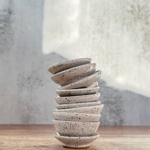 May include: A stack of small, white ceramic bowls with a speckled glaze. The bowls are stacked in a staggered pattern, with the largest bowl at the bottom and the smallest bowl at the top.