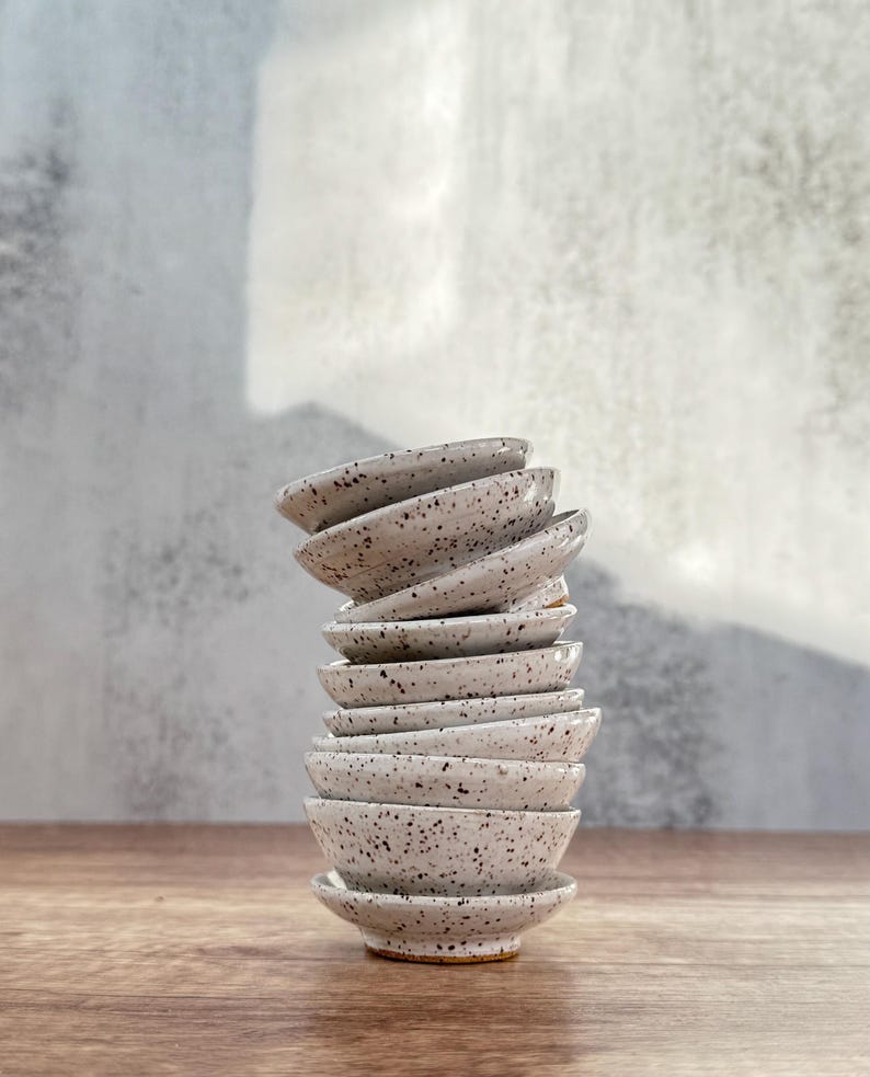 May include: A stack of small, white ceramic bowls with a speckled glaze. The bowls are stacked in a staggered pattern, with the largest bowl at the bottom and the smallest bowl at the top.