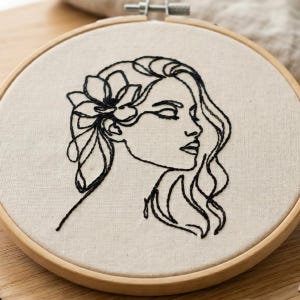 May include: An embroidery hoop with a black line art design of a woman's profile, flowing hair, and a flower. The design is stitched on a beige fabric, set within a wooden hoop.