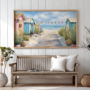 Op de afbeelding: Een ingelijst schilderij toont een strandscène met kleurrijke strandhuisjes, een houten loopbrug en de oceaan. Het kunstwerk wordt tentoongesteld boven een houten bank met witte kussens. Decoratieve planten en een vaas staan op de voorgrond.