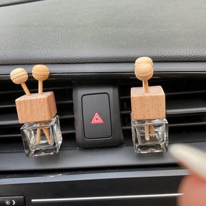 May include: Two car air fresheners with wooden tops and brown wooden sticks. The air fresheners are in clear glass bottles and are placed in the air vents of a car.