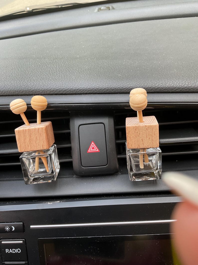 May include: Two car air fresheners with wooden tops and brown wooden sticks. The air fresheners are in clear glass bottles and are placed in the air vents of a car.
