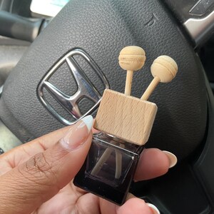 May include: A small, square, dark brown glass bottle with a wooden top and two wooden sticks. The bottle is filled with a clear liquid and the sticks are partially submerged in the liquid. The bottle is being held in a person's hand.