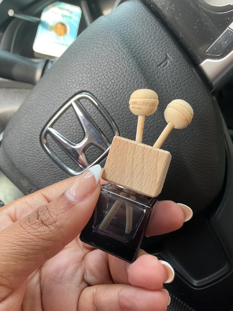 May include: A small, square, dark brown glass bottle with a wooden top and two wooden sticks. The bottle is filled with a clear liquid and the sticks are partially submerged in the liquid. The bottle is being held in a person's hand.