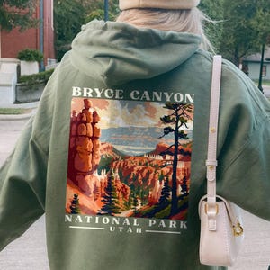 May include: Military green hoodie with a Bryce Canyon National Park graphic on the back. The design features a landscape illustration with the text "BRYCE CANYON" and "NATIONAL PARK UTAH". The hoodie has a drawstring hood.