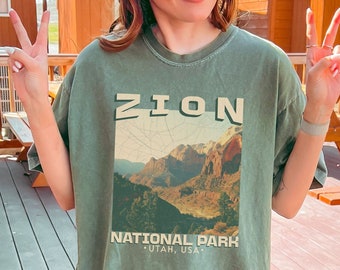 May include: A moss green t-shirt with a graphic print of Zion National Park, Utah, USA. The text "Zion National Park" and "Utah, USA" are printed in white on the shirt. The shirt is oversized and has a relaxed fit.