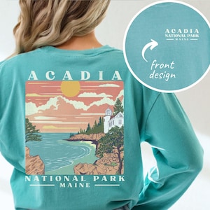 May include: Seafoam green long-sleeve shirt featuring a scenic Acadia National Park design on the back. The design includes a coastal landscape with a lighthouse, sun, and the text "ACADIA NATIONAL PARK MAINE." A smaller design is on the front.