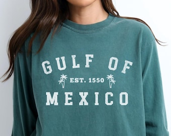 May include: A teal blue long-sleeved t-shirt with the text "GULF OF EST. 1550 MEXICO" in white. The shirt has a distressed look and is worn by a person.