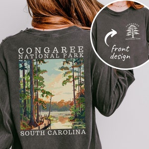 May include: A dark gray long-sleeve t-shirt with a vintage-style illustration of Congaree National Park in South Carolina. The back of the shirt features the park's name and location. The front of the shirt has a small logo with a tree and the words "Congaree National Park".