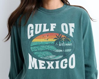 May include: A teal blue long-sleeved t-shirt with a vintage-style graphic that says "GULF OF MEXICO" with a sunset and palm trees.