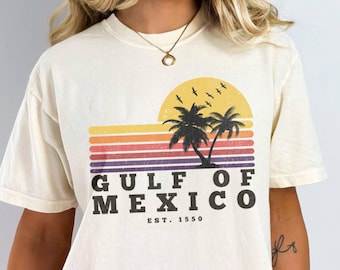 May include: Ivory short-sleeved t-shirt with a retro-style graphic design featuring palm trees, a sunset, and the text "GULF OF MEXICO EST. 1550".
