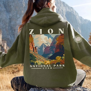 May include: Military green hoodie with a Zion National Park graphic on the back. The design features the text "ZION" above a colorful landscape illustration and the text "NATIONAL PARK UTAH". The hoodie is a comfortable, casual style.
