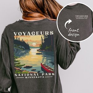 May include: Dark grey long-sleeve shirt featuring a scenic illustration of Voyageurs National Park, Minnesota. The design includes a lake, trees, a canoe, and the text "VOYAGEURS NATIONAL PARK MINNESOTA". A smaller design is on the front. From Red Prairie Goods & Gifts.