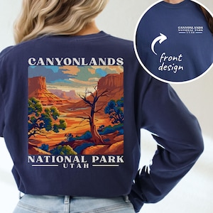 May include: Midnight blue long-sleeve shirt featuring a Canyonlands National Park Utah graphic on the back. The design includes a landscape illustration with the text "CANYONLANDS NATIONAL PARK UTAH". A smaller front design is also shown.