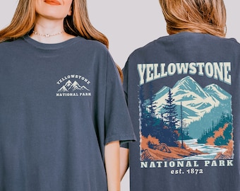 May include: Denim-colored t-shirt with a front and back design. The front features the text "Yellowstone National Park" above a mountain graphic. The back displays a larger graphic of a mountain landscape with the text "Yellowstone National Park est. 1872".