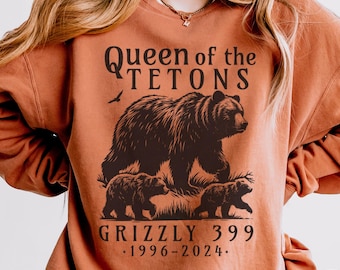 Può includere: Una felpa marrone con una stampa grafica di un orso e cuccioli. Il testo sulla felpa recita "Queen of the Tetons Grizzly 399 1996-2024."