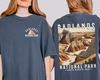 May include: A navy blue t-shirt with a graphic of Badlands National Park on the back. The front of the shirt has a smaller graphic of the park with the text "Badlands National Park".