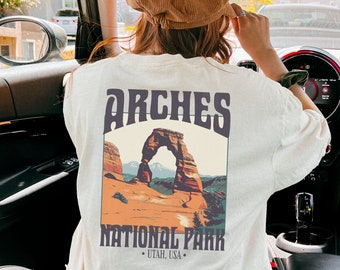 Op de afbeelding: Een wit T-shirt met een vintage-stijl grafische afbeelding van Arches National Park in Utah. De afbeelding toont een rode rotsboog met een bergketen op de achtergrond. De tekst "Arches National Park" is onder de afbeelding gedrukt.