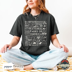 May include: Dark gray t-shirt featuring a white graphic design of the words "National Parks of America" surrounded by park names and illustrations. The shirt is a comfortable, casual style.