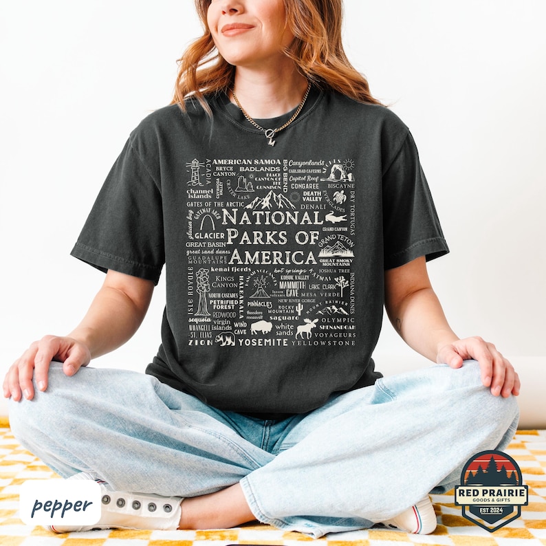May include: Dark gray t-shirt featuring a white graphic design of the words "National Parks of America" surrounded by park names and illustrations. The shirt is a comfortable, casual style.