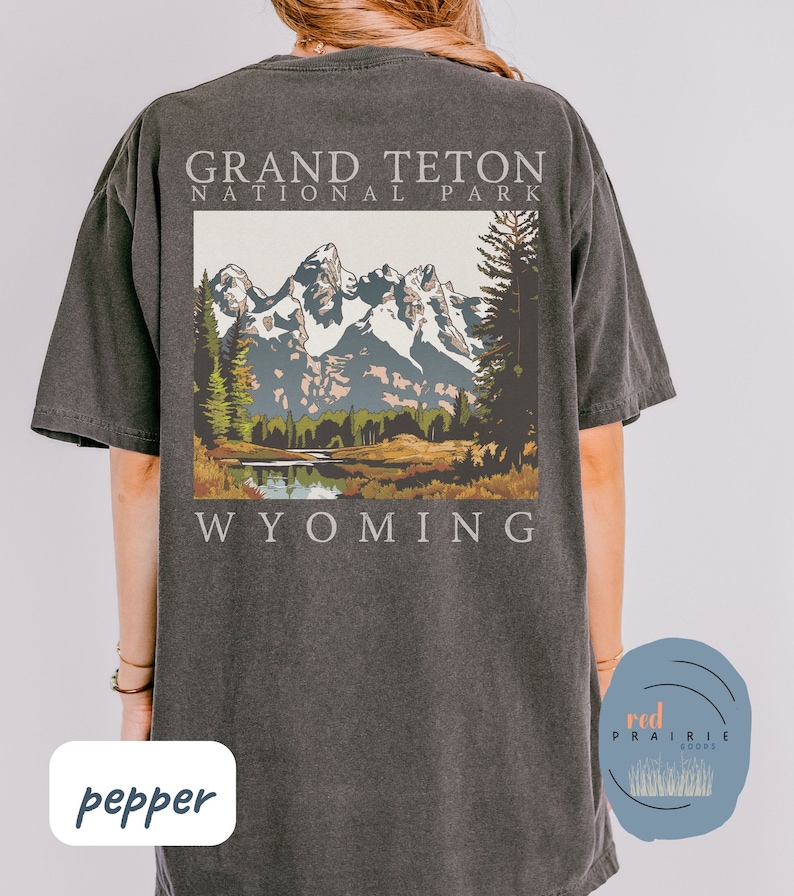 May include: A dark gray t-shirt with a vintage-style illustration of the Grand Teton National Park in Wyoming. The illustration features a mountain range, trees, and a lake. The text "Grand Teton National Park" and "Wyoming" are printed below the illustration.