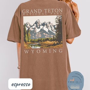 May include: Espresso brown t-shirt with a graphic of the Grand Teton National Park in Wyoming. The graphic features a mountain range with snow-capped peaks, a lake, and trees. The text "Grand Teton National Park" and "Wyoming" are printed on the shirt.