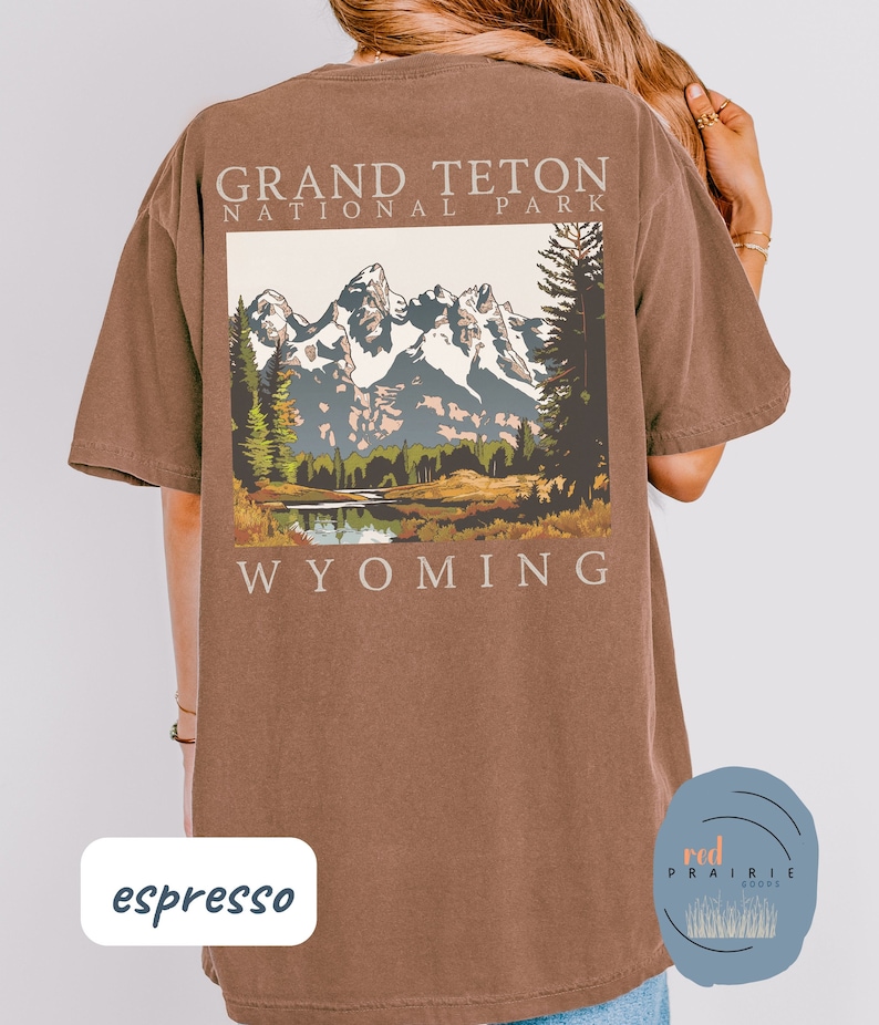 May include: Espresso brown t-shirt with a graphic of the Grand Teton National Park in Wyoming. The graphic features a mountain range with snow-capped peaks, a lake, and trees. The text "Grand Teton National Park" and "Wyoming" are printed on the shirt.