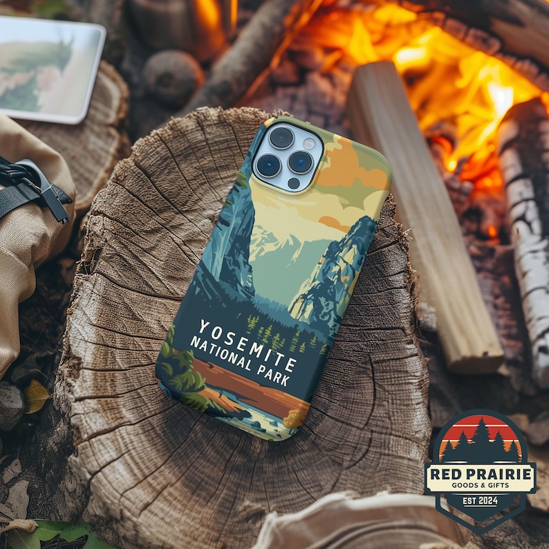 May include: A phone case featuring a Yosemite National Park landscape design with mountains, trees, and a river. The case has a dark blue background with the text "YOSEMITE NATIONAL PARK". The case is on a wooden surface.