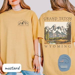 May include: Two mustard yellow t-shirts with a graphic design of the Grand Teton National Park in Wyoming. The front of one shirt has a small graphic of the mountains and the text "Grand Teton National Park". The back of the other shirt has a larger graphic of the mountains and the text "Grand Teton National Park Wyoming".