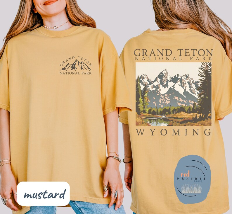 May include: Two mustard yellow t-shirts with a graphic design of the Grand Teton National Park in Wyoming. The front of one shirt has a small graphic of the mountains and the text "Grand Teton National Park". The back of the other shirt has a larger graphic of the mountains and the text "Grand Teton National Park Wyoming".