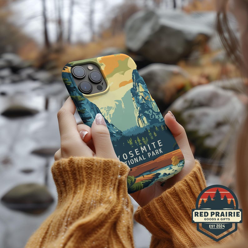 May include: A phone case featuring a landscape design of Yosemite National Park. The case has a colorful illustration of mountains, trees, and a river, with the text "YOSEMITE NATIONAL PARK". The case is held in a person's hands.