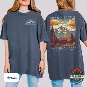 May include: Denim blue t-shirt with a front graphic of the Grand Teton National Park logo and a back graphic of a mountain landscape scene. The back graphic includes the text "GRAND TETON NATIONAL PARK WYOMING EST. 1929".