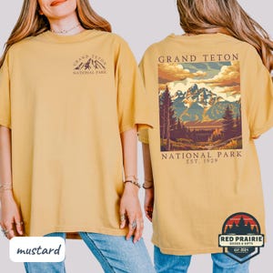 May include: Mustard-colored t-shirt featuring a front and back design. The front displays "Grand Teton National Park" with a mountain graphic. The back showcases a landscape scene of mountains and trees, with text "Grand Teton National Park, Est. 1929".
