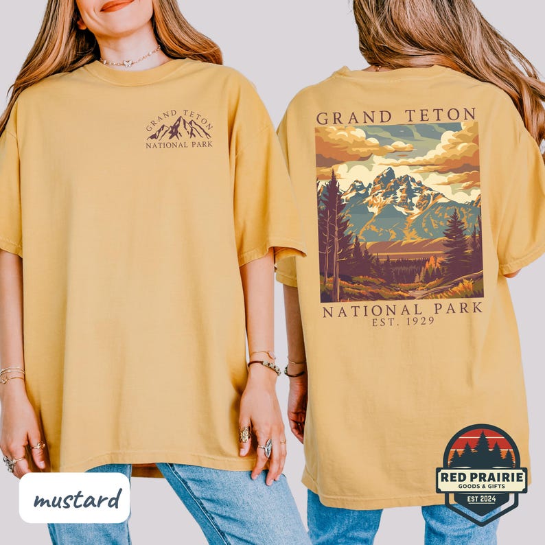 May include: Mustard-colored t-shirt featuring a front and back design. The front displays "Grand Teton National Park" with a mountain graphic. The back showcases a landscape scene of mountains and trees, with text "Grand Teton National Park, Est. 1929".