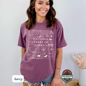 May include: Berry-colored t-shirt featuring a graphic design listing various National Parks of America. The design includes illustrations and text, with the words "National Parks of America" prominently displayed. The shirt also has the word "berry" printed on it.