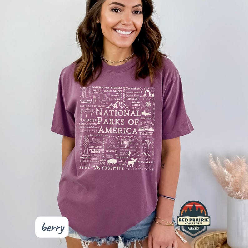 May include: Berry-colored t-shirt featuring a graphic design listing various National Parks of America. The design includes illustrations and text, with the words "National Parks of America" prominently displayed. The shirt also has the word "berry" printed on it.