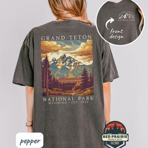 May include: A dark gray t-shirt featuring a colorful graphic of the Grand Teton National Park in Wyoming. The design includes mountains, trees, and the text "GRAND TETON NATIONAL PARK WYOMING EST. 1929". A smaller design is on the front.