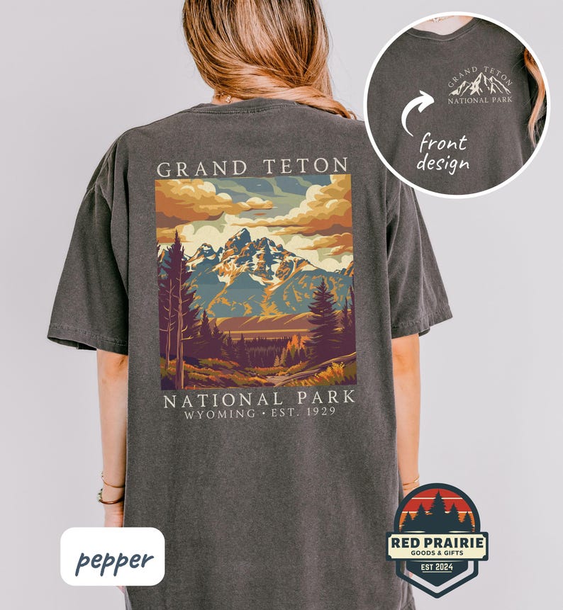 May include: A dark gray t-shirt featuring a colorful graphic of the Grand Teton National Park in Wyoming. The design includes mountains, trees, and the text "GRAND TETON NATIONAL PARK WYOMING EST. 1929". A smaller design is on the front.
