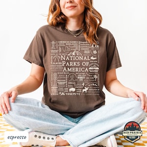 May include: Espresso-colored t-shirt featuring a graphic of National Parks of America. The design includes park names and illustrations in white. The shirt is worn by a person sitting cross-legged.