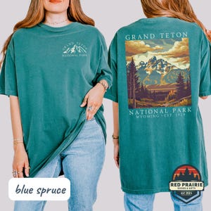 May include: Blue spruce t-shirt featuring a Grand Teton National Park design. The front displays the park name and mountain graphic, while the back showcases a scenic landscape with mountains, trees, and the park's name. Includes the text "Wyoming Est. 1929".