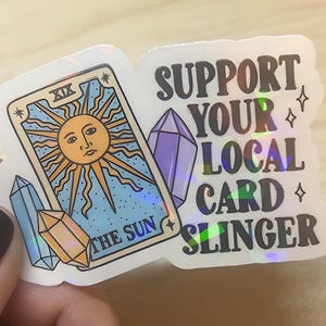 May include: A sticker featuring a tarot card design with a sun illustration and the text "THE SUN". The sticker also includes the text "SUPPORT YOUR LOCAL CARD SLINGER" and crystal graphics.