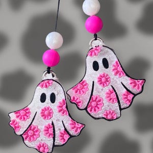 May include: Two ghost-shaped ornaments featuring a glittery silver finish with raised pink floral patterns and black oval eyes. Each ghost hangs from a black cord accented with one white bead and one bright pink bead, creating a decorative pair.