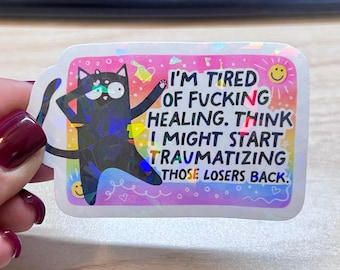 May include: A rectangular holographic sticker featuring a black cat with wide eyes and the text I'M TIRED OF FUCKING HEALING. THINK I MIGHT START TRAUMATIZING THOSE LOSERS BACK. The background is a vibrant pink and orange gradient with small yellow smiley faces.